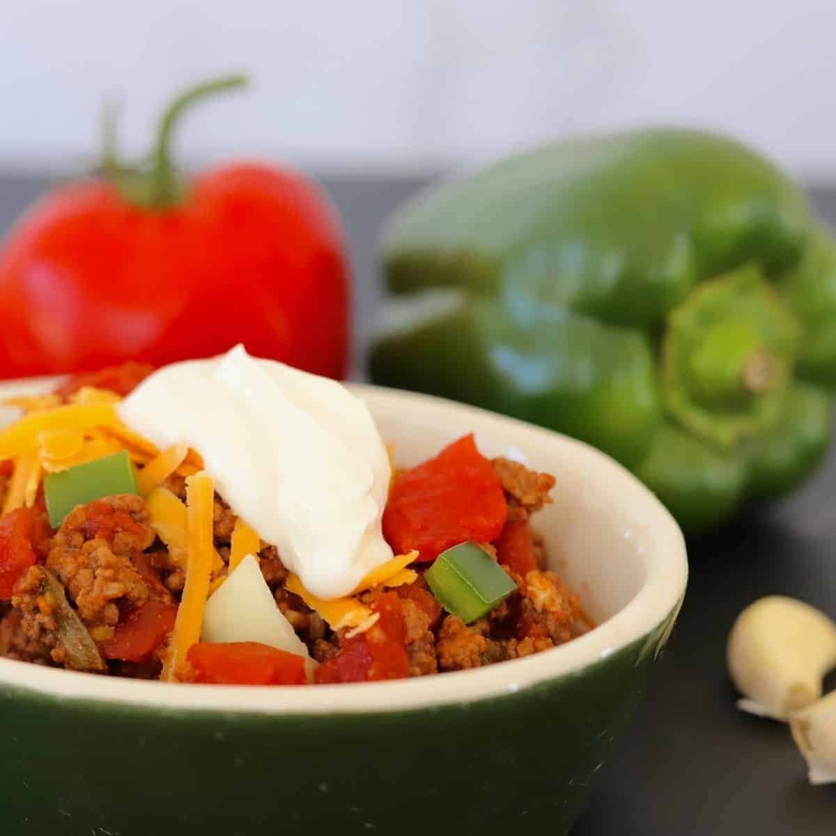 Bowl full of chili next to a tomato and green pepper, topped with shredded cheese and a dollop of sour cream.