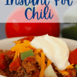 Bowl full of chili next to a tomato and green pepper, topped with shredded cheese and a dollop of sour cream.