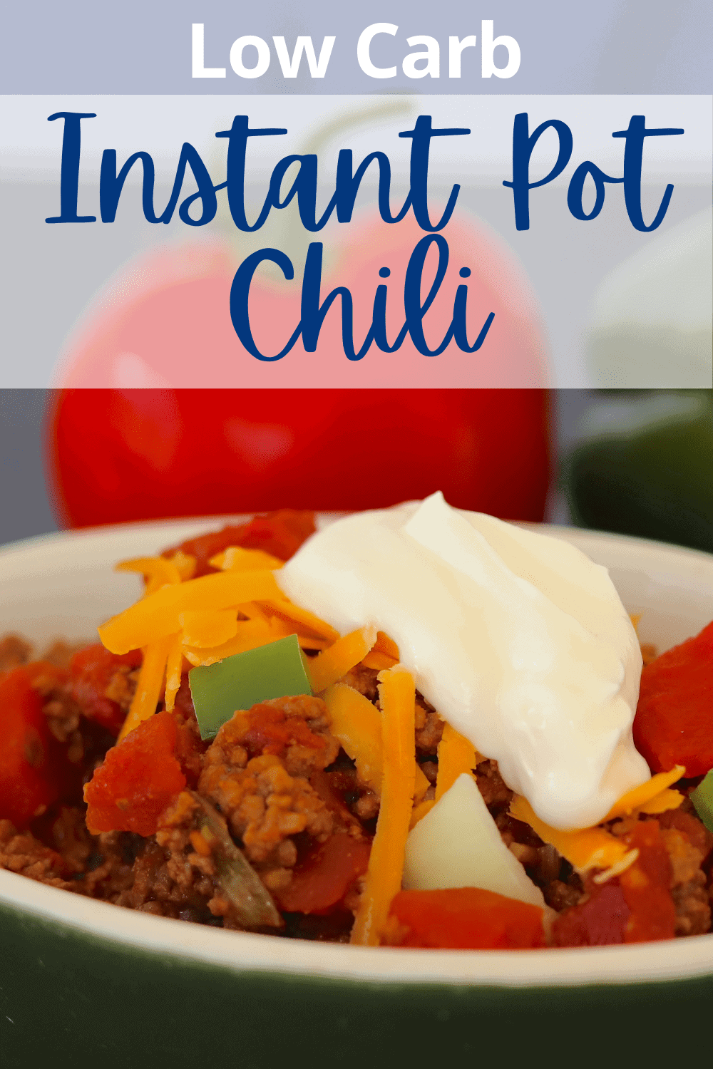 Bowl full of chili next to a tomato and green pepper, topped with shredded cheese and a dollop of sour cream.