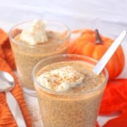 Two glasses filled with pumpkin chia pudding topped with whipped cream and a sprinkle of pumpkin spice with fall foliage in the background.