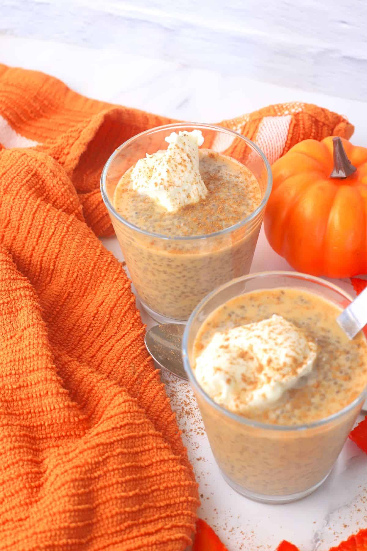 Two glasses filled with pumpkin chia pudding topped with whipped cream and a sprinkle of pumpkin spice with fall foliage in the background.