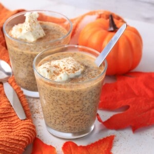 Two glasses filled with pumpkin chia pudding topped with whipped cream and a sprinkle of pumpkin spice with fall foliage in the background.