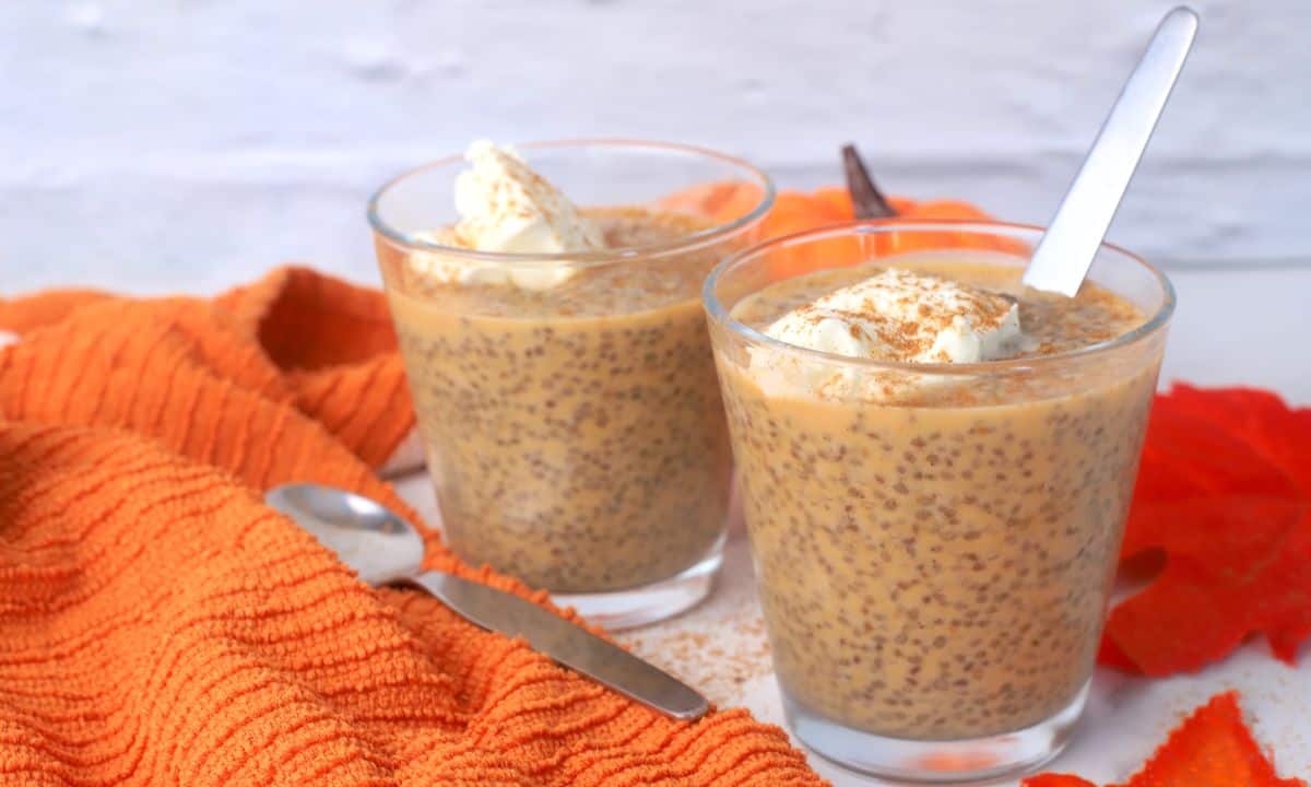 Two glasses filled with pumpkin chia pudding topped with whipped cream and a sprinkle of pumpkin spice with fall foliage in the background.