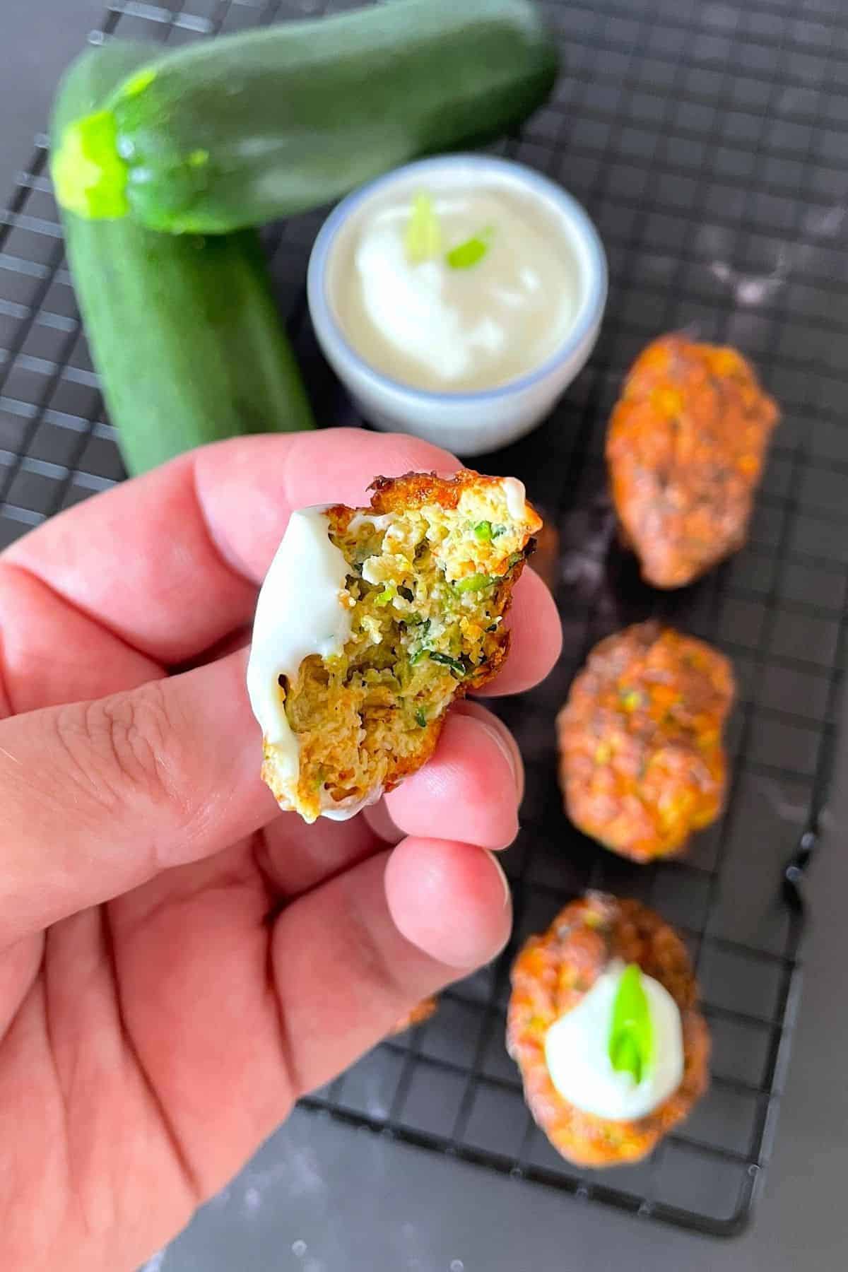 holding half of a fritter topped with sour cream above more fritters on a cooling rack with dipping bowl and 2 raw zucchinis