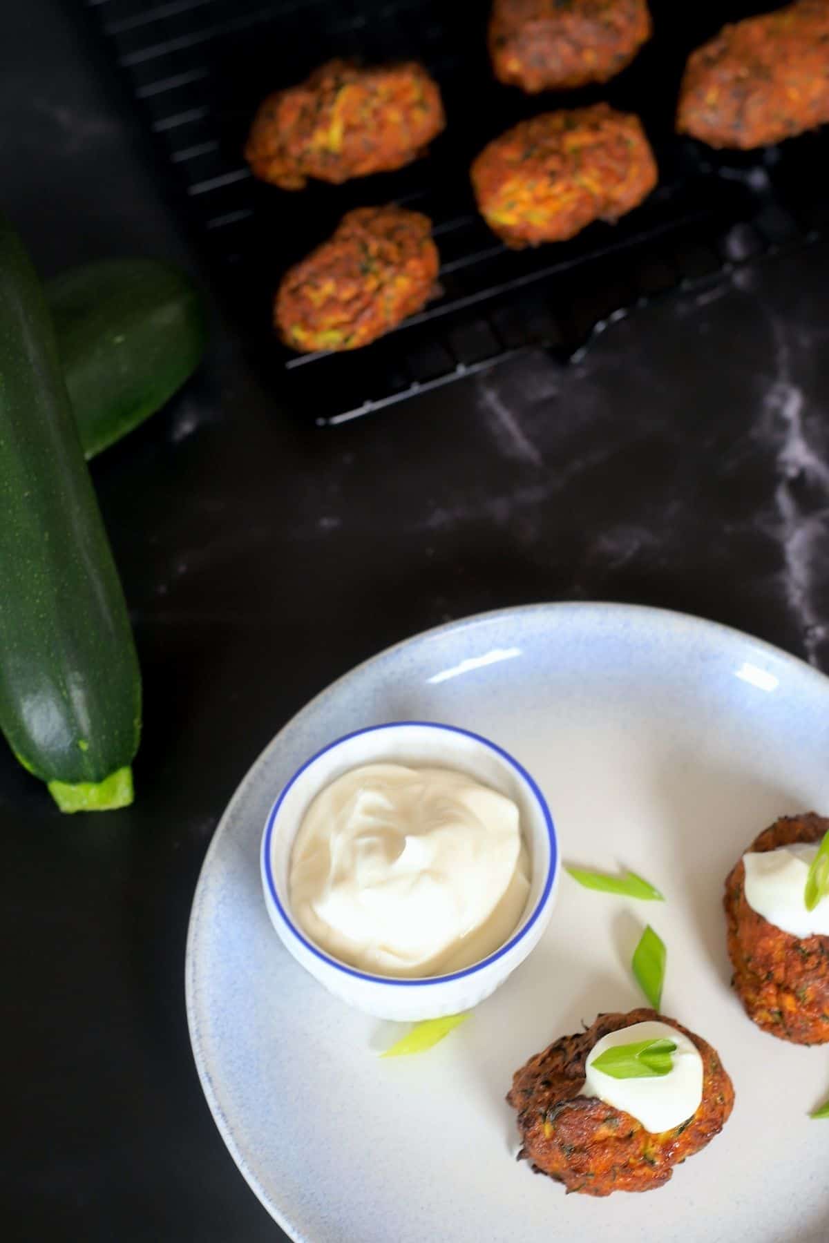 zucchini fritters on a cooling rack topped with sour cream and onion 2 on a plate with dipping bowl and 2 zucchinis