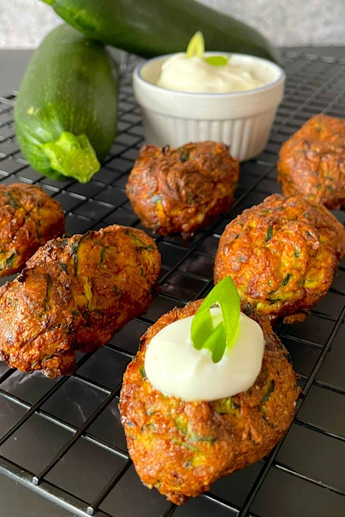 zucchini fritters on a cooling rack topped with sour cream and onion with a dipping bowl and two raw zucchinis