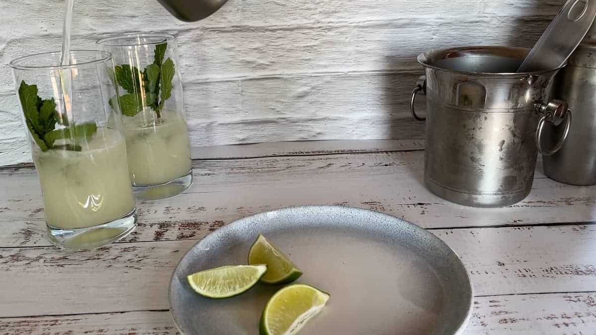 Pouring from shaker into highball glass with a side plate of lime wedges and an ice bucket