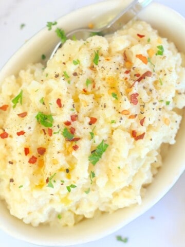cauliflower mash in a bowl with a spoon topped with chili flakes, olive oil and parsley