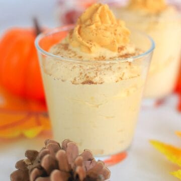 two pumpkin mousse in a glass topped with whipped cream next to acorns and a pumpkin