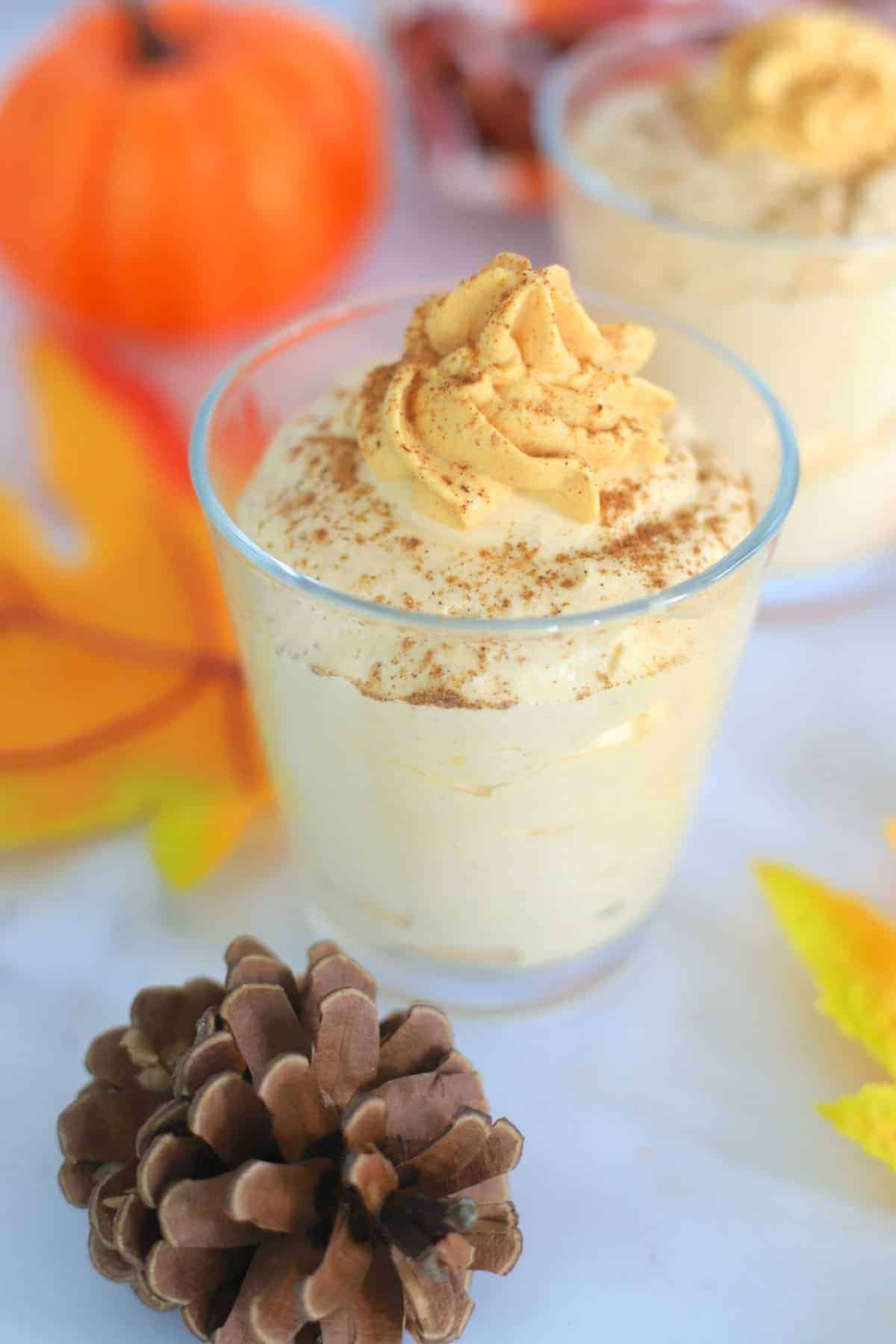 two pumpkin mousse in a glass topped with whipped cream next to acorns and a pumpkin