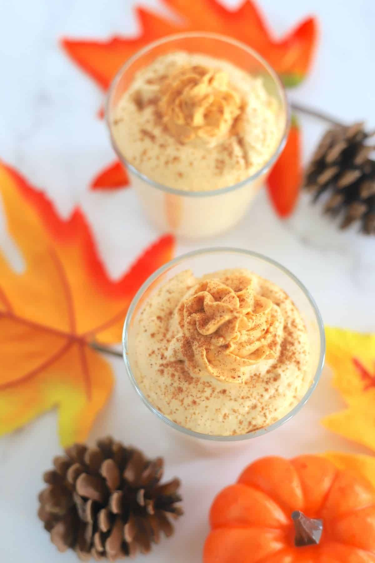 two pumpkin mousse in a glass topped with whipped cream next to acorns and a pumpkin