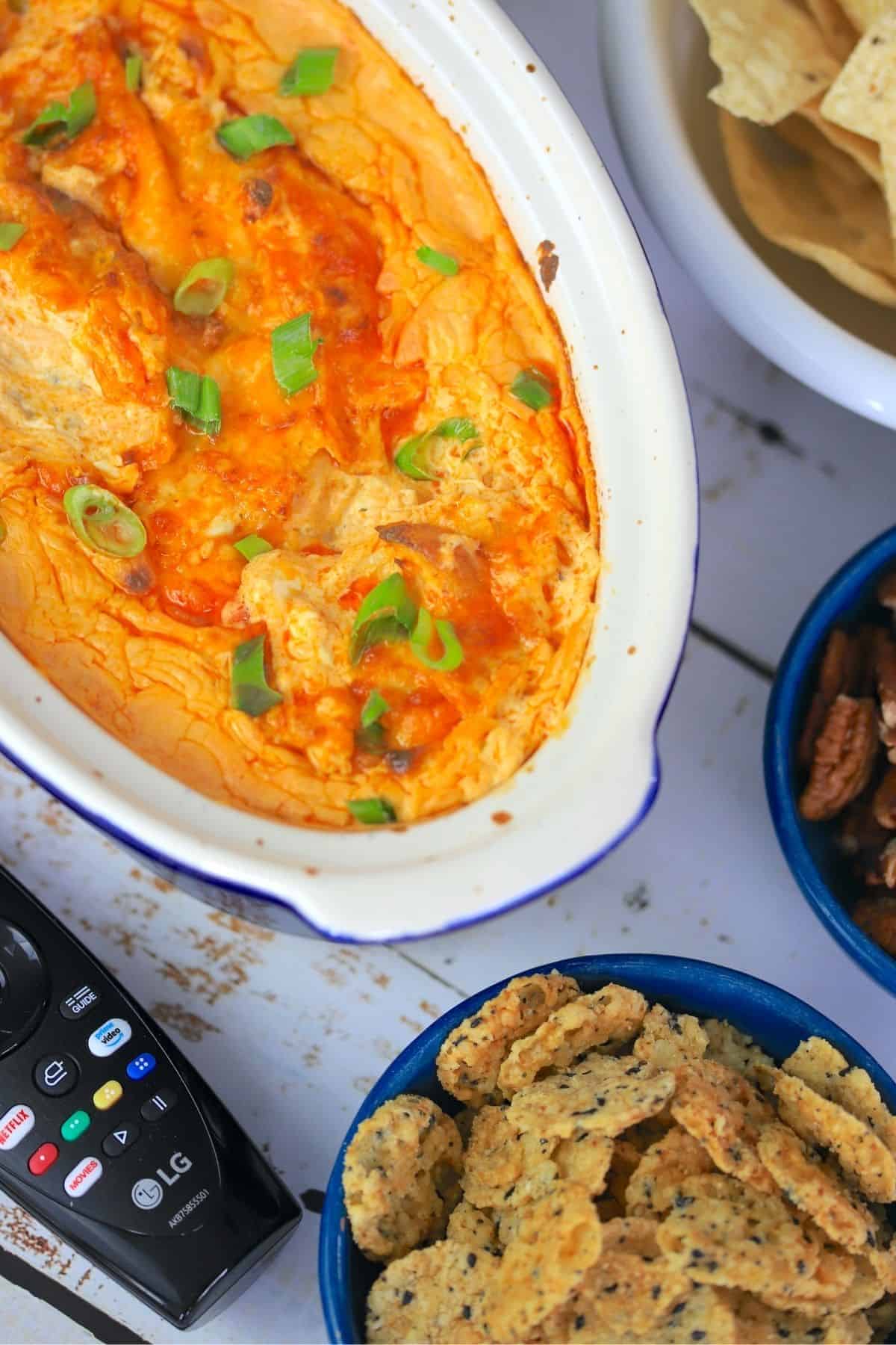 casserole of buffalo dip with bowls of nacho chips pecans and cheese crackers and a tv remote