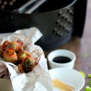 brussel sprouts wrapped in bacon with a toothpick inserted next to air fryer basket, toothpicks and small bowl of soy sauce