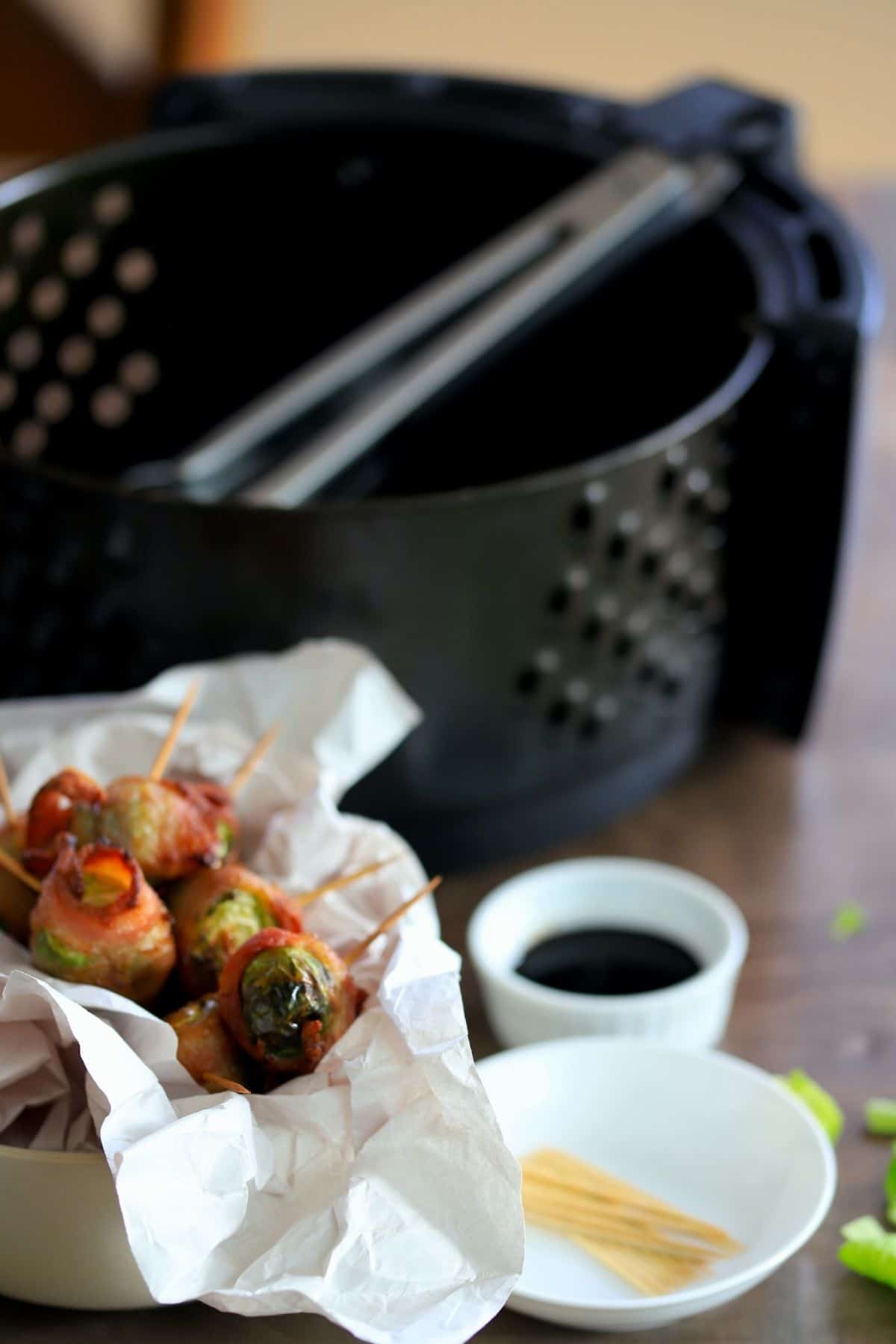 brussel sprouts wrapped in bacon with a toothpick inserted next to air fryer basket, toothpicks and small bowl of soy sauce