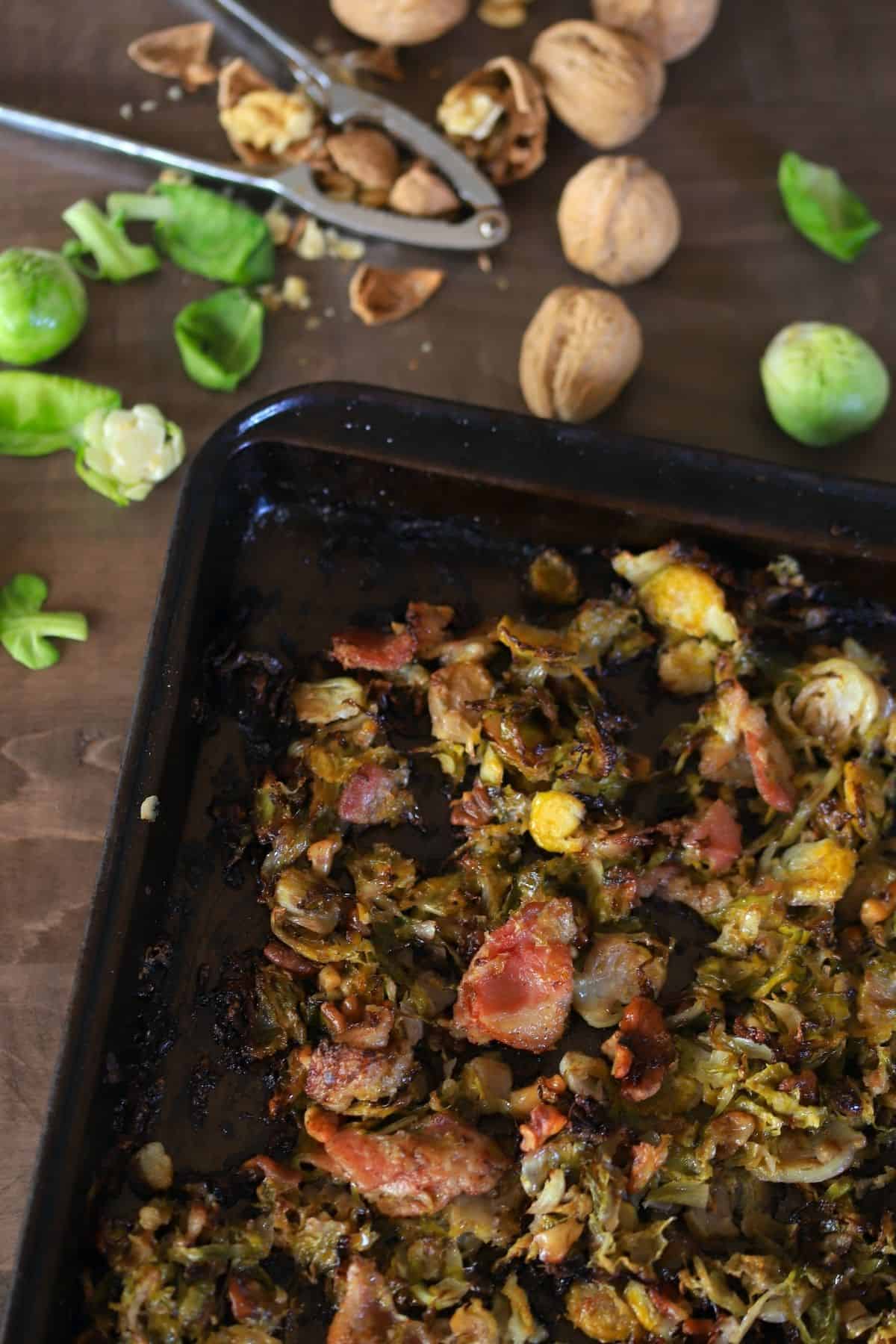 baked brussel sprouts on a baking sheet next to cracked walnuts with nutcracker and some brussel sprout leaves