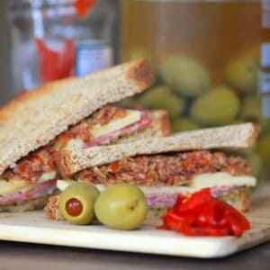 deli sandwich topped with muffaletta, next to 2 olives some roasted red peppers and a jar of olives