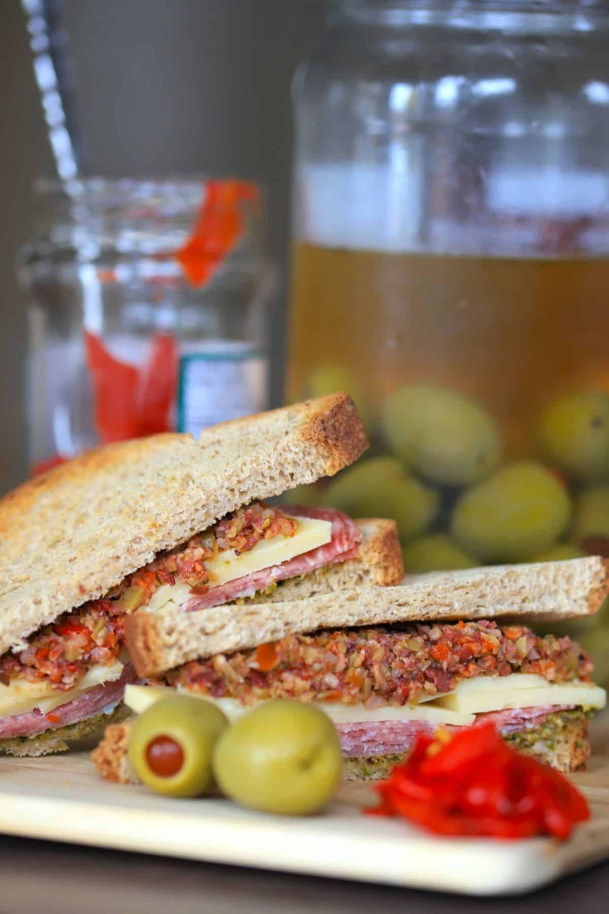 deli sandwich topped with muffaletta, next to 2 olives some roasted red peppers and a jar of olives