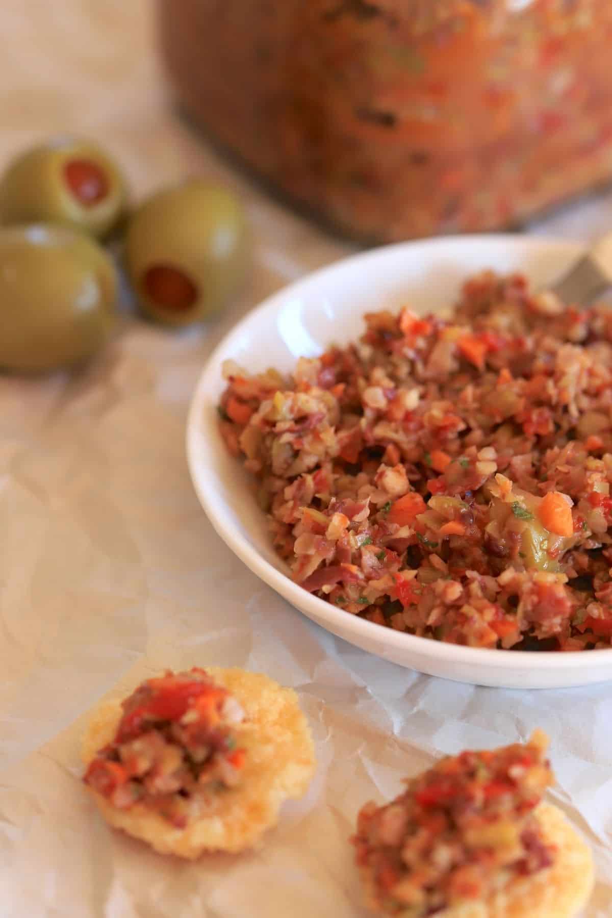 muffaletta spread in a small bowl next to 3 olives a jar and two cheese crackers topped with spread