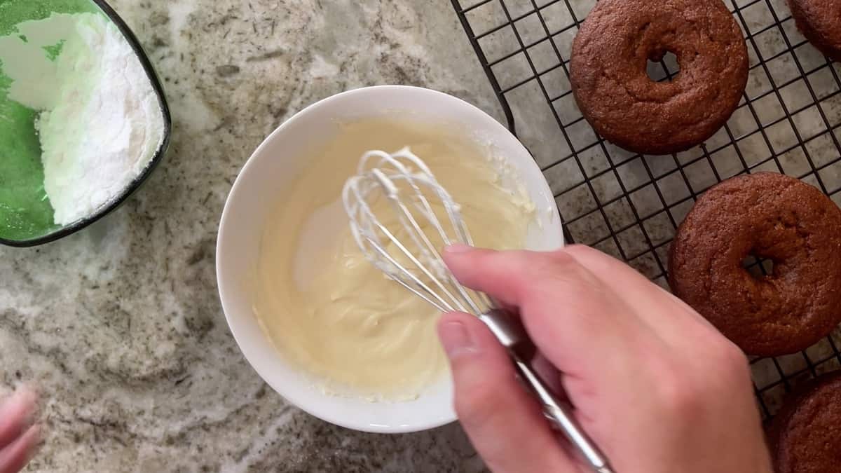 mixing white glaze in a white bowl next to donuts on a cooling rack