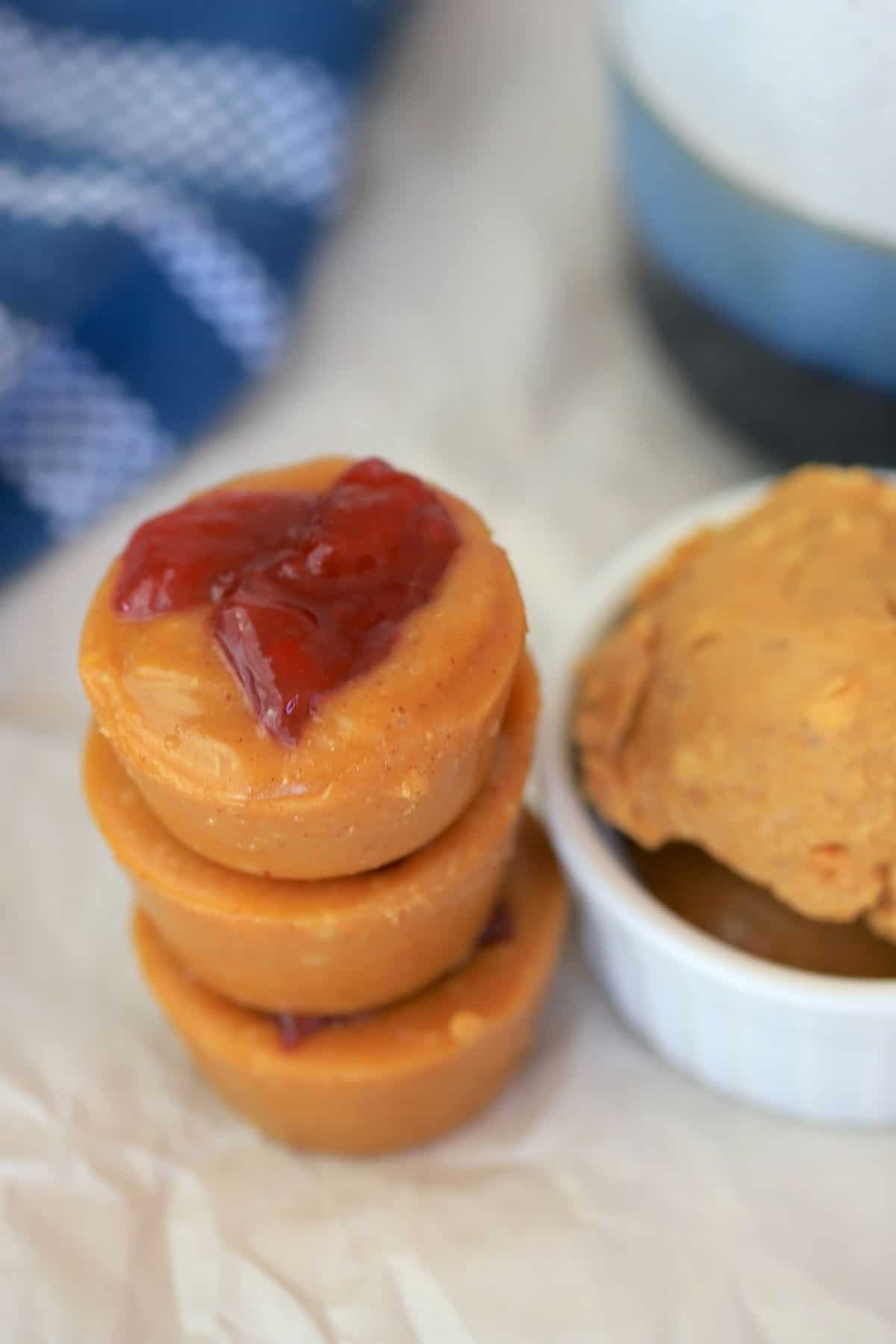 Sugar free peanut butter fudge bombs stacked on top of each other next to a bowl with peanut butter and a blue kitchen towel.