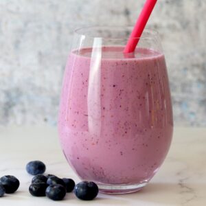 low carb berry smoothie in a clear glass with a pink straw next to blueberries.