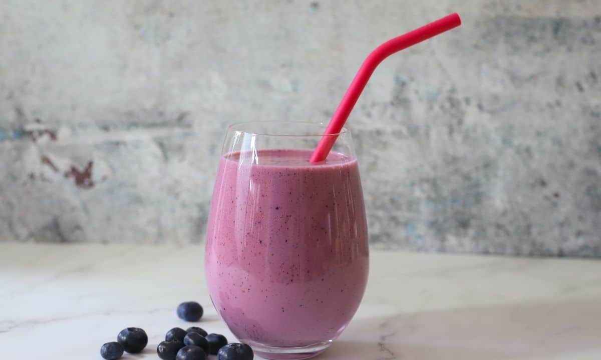 low carb berry smoothie in a clear glass with a pink straw next to blueberries.