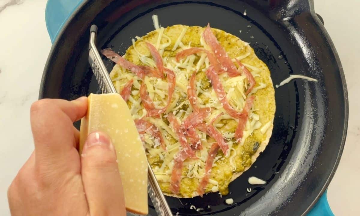 grating parmesan on a tortilla on a skillet topped with pesto, cheese and salami.