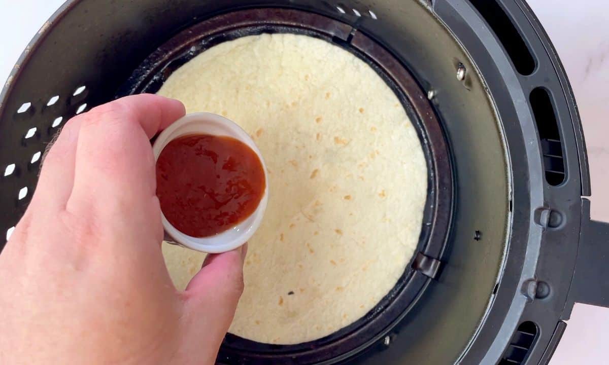 adding sauce to the tortilla in the air fryer basket.