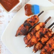 bacon wrapped chicken skewers on a white plate with a side bowl of bbq sauce.