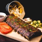 racks of ribs on a cutting board with tomatoes, pickles and fries.