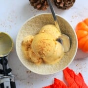 a bowl of keto pumpkin ice cream with a spoon next to ice cream scoop, acorns and a small pumpkin.