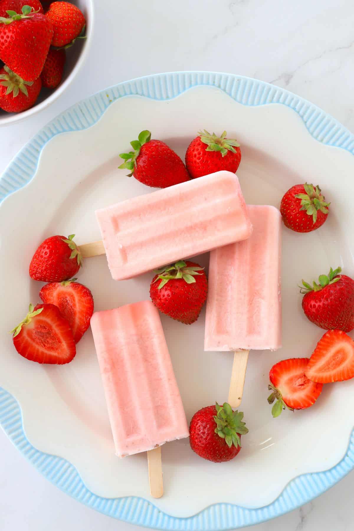 sugar free strawberry ice cream popsicles on a plate with fresh strawberries and a white bowl with fresh strawberries.