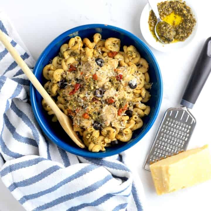 pesto chicken on pasta in a blue dish next to parmesan grater and a small bowl of pesto.