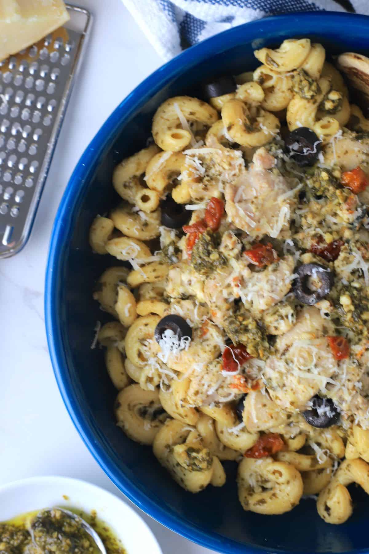 pesto chicken on pasta in a blue dish next to parmesan grater and a small bowl of pesto.