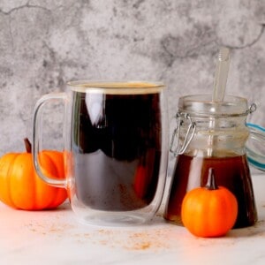 cup of black coffee with jar of sugar free pumpkin syrup on the side with pumpkin ornaments.