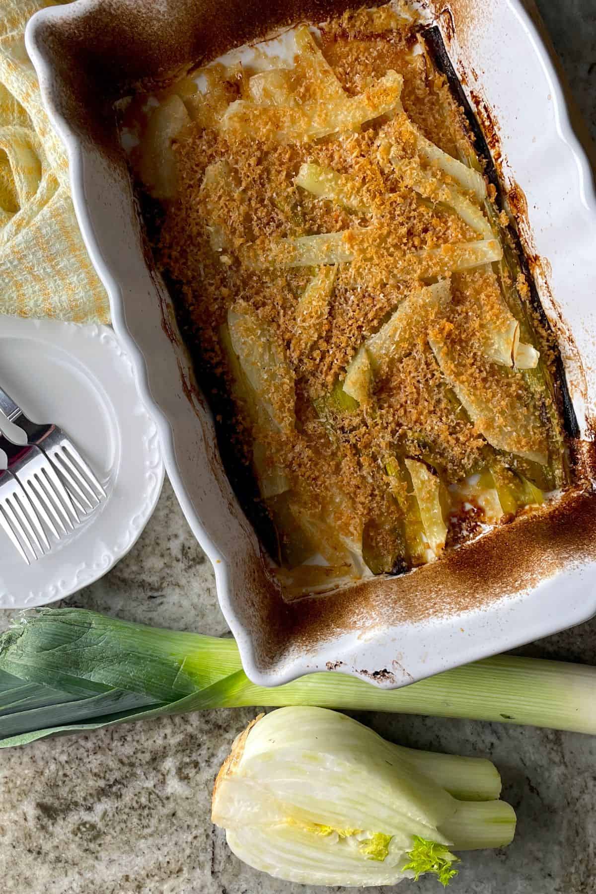 braised fennel and leeks in a white casserole dish topped with a parmesan crust with a plate on the side and a raw fennel and leek.