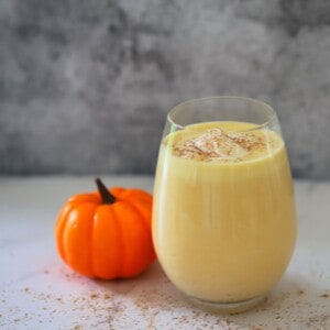 a glass filled with keto pumpkin smoothie with a dollop of whip cream on top and sprinkles of pumpkin spice next to a mini pumpkin ornament.