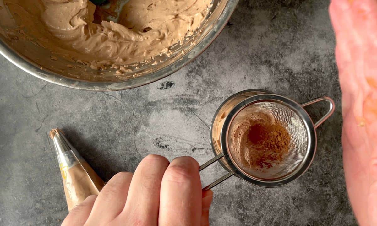 sprinkling cocoa powder into the tumbler next to a piping bag and a bowl.