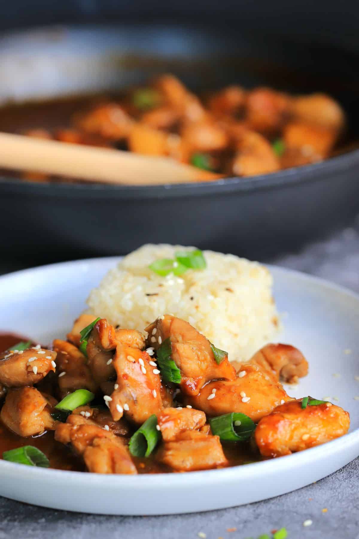 a dish with keto bourbon chicken and a side of cauliflower rice topped with green onions and sesame seeds.