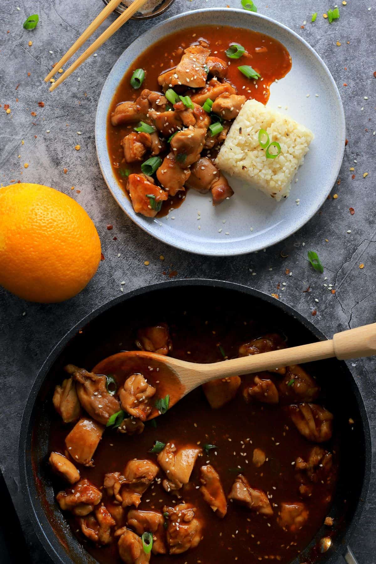 a skillet with keto bourbon chicken and a wooden spoon next to a dish with the bourbon chicken and cauliflower rice.