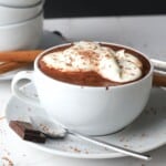 white cup on a saucer filled with hot chocolate, dollops of whip cream and a piece of chocolate on the side with cinnamon sticks.