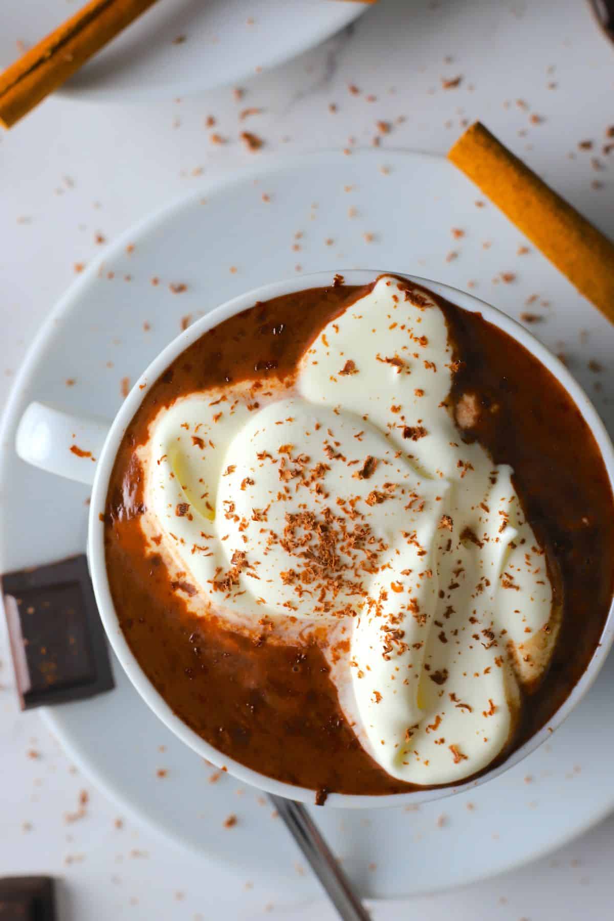 white cup on a saucer filled with hot chocolate, dollops of whip cream and a piece of chocolate on the side with cinnamon sticks.