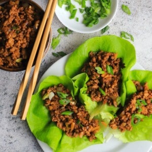 three chicken lettuce wraps on a white dish with a bowl of chicken, two chopsticks and a side dish with sliced green onions.