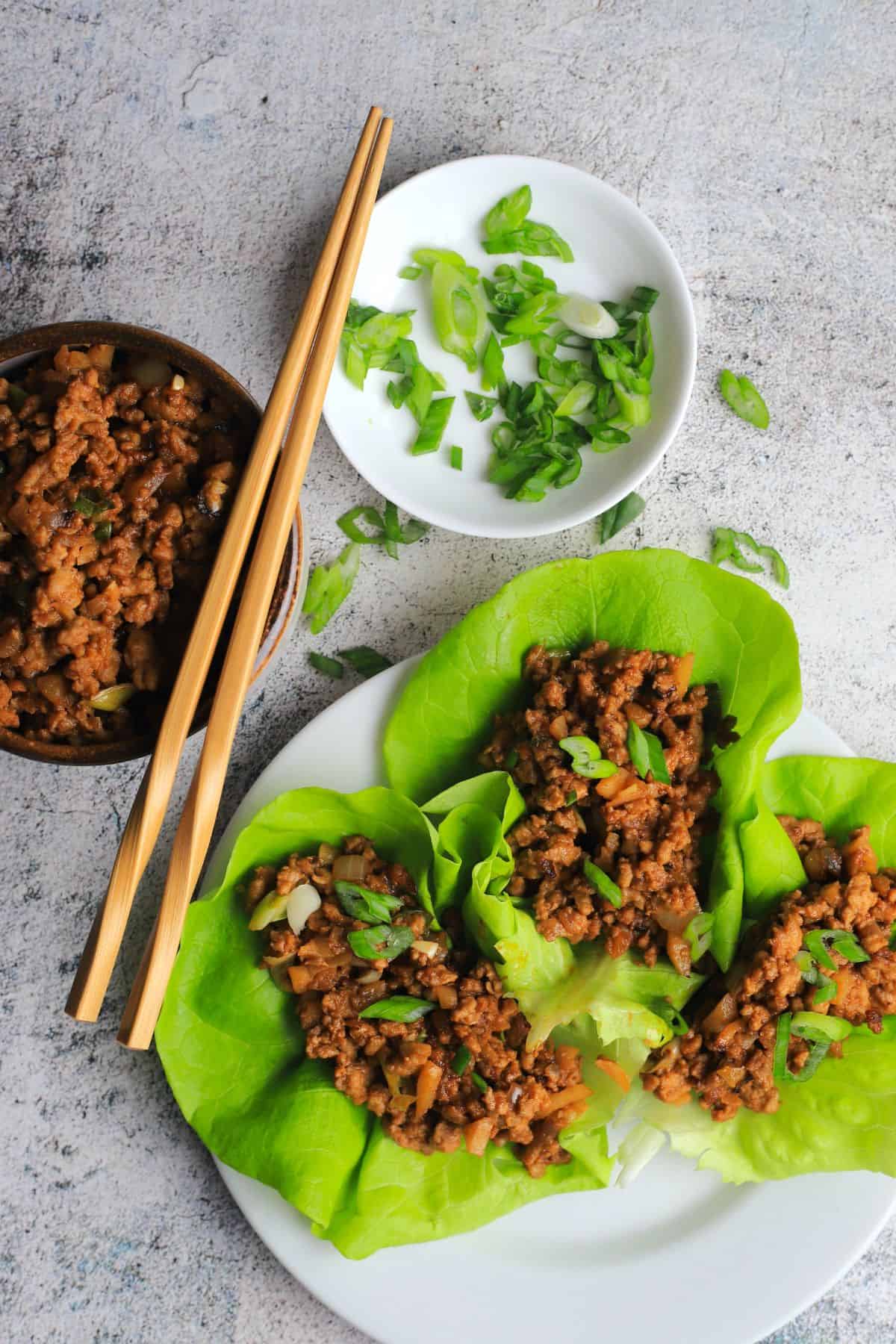 three chicken lettuce wraps on a white dish with a bowl of chicken, two chopsticks and a side dish with sliced green onions.