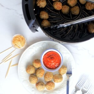 meatballs in the air fryer basket with tongs next to a white plate with air fried meatballs and a small bowl of ketchup.