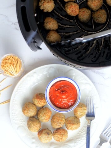 meatballs in the air fryer basket with tongs next to a white plate with air fried meatballs and a small bowl of ketchup.