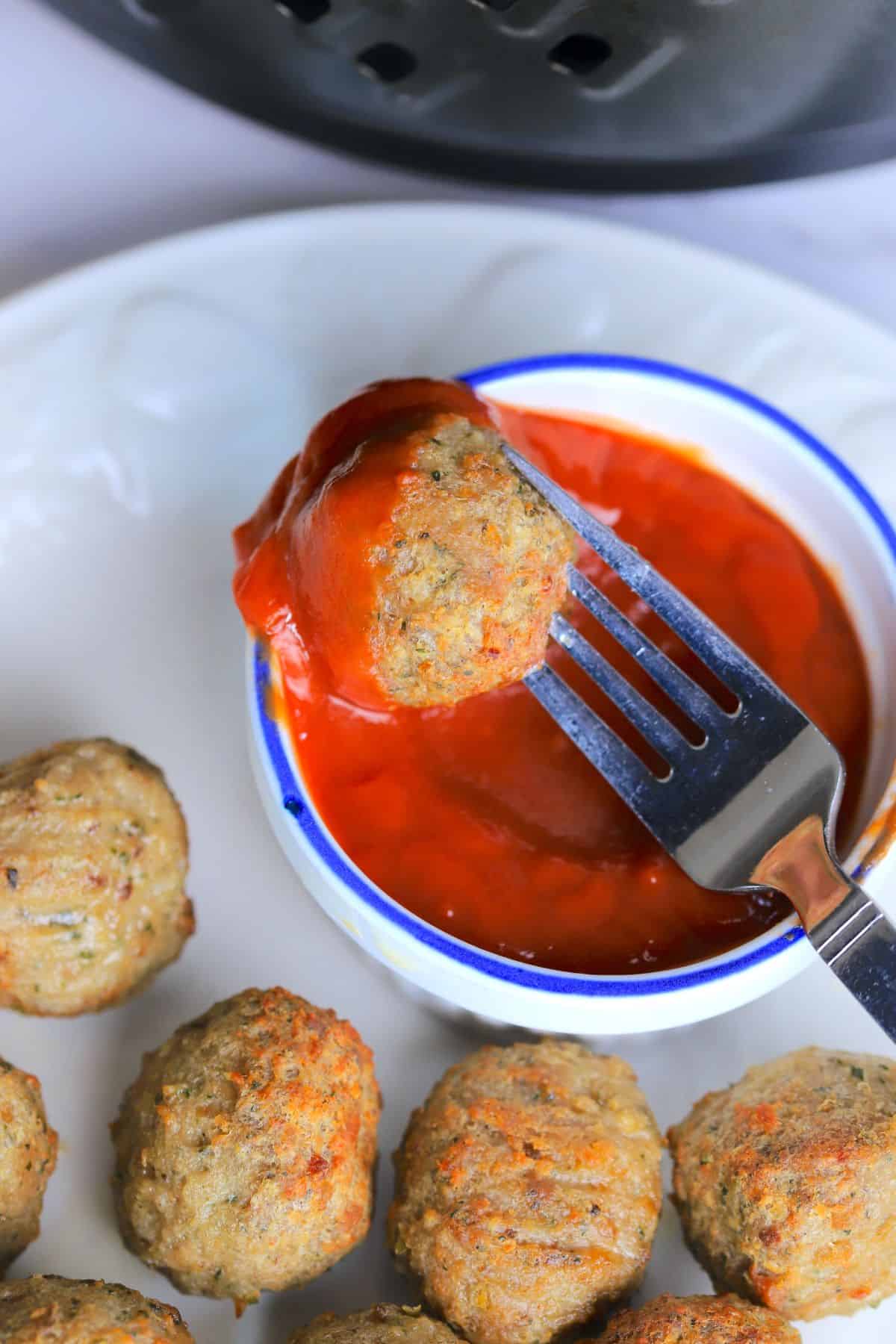 meatballs in the air fryer basket with tongs next to a white plate with air fried meatballs and a small bowl of ketchup.