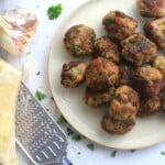 Fried meatballs on a plate next to a garlic head, a block of cheese, and a cheese grater.