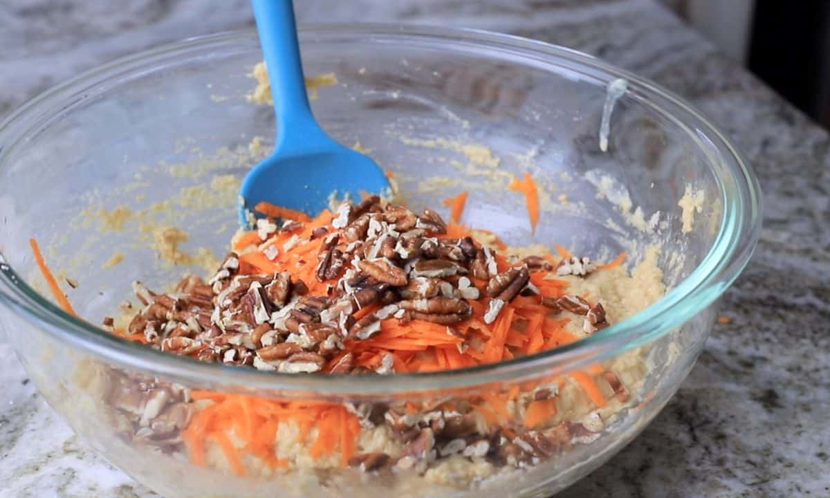 grated carrot and toasted pecans on top of the cake batter in a glass bowl with a blue rubber spatula.