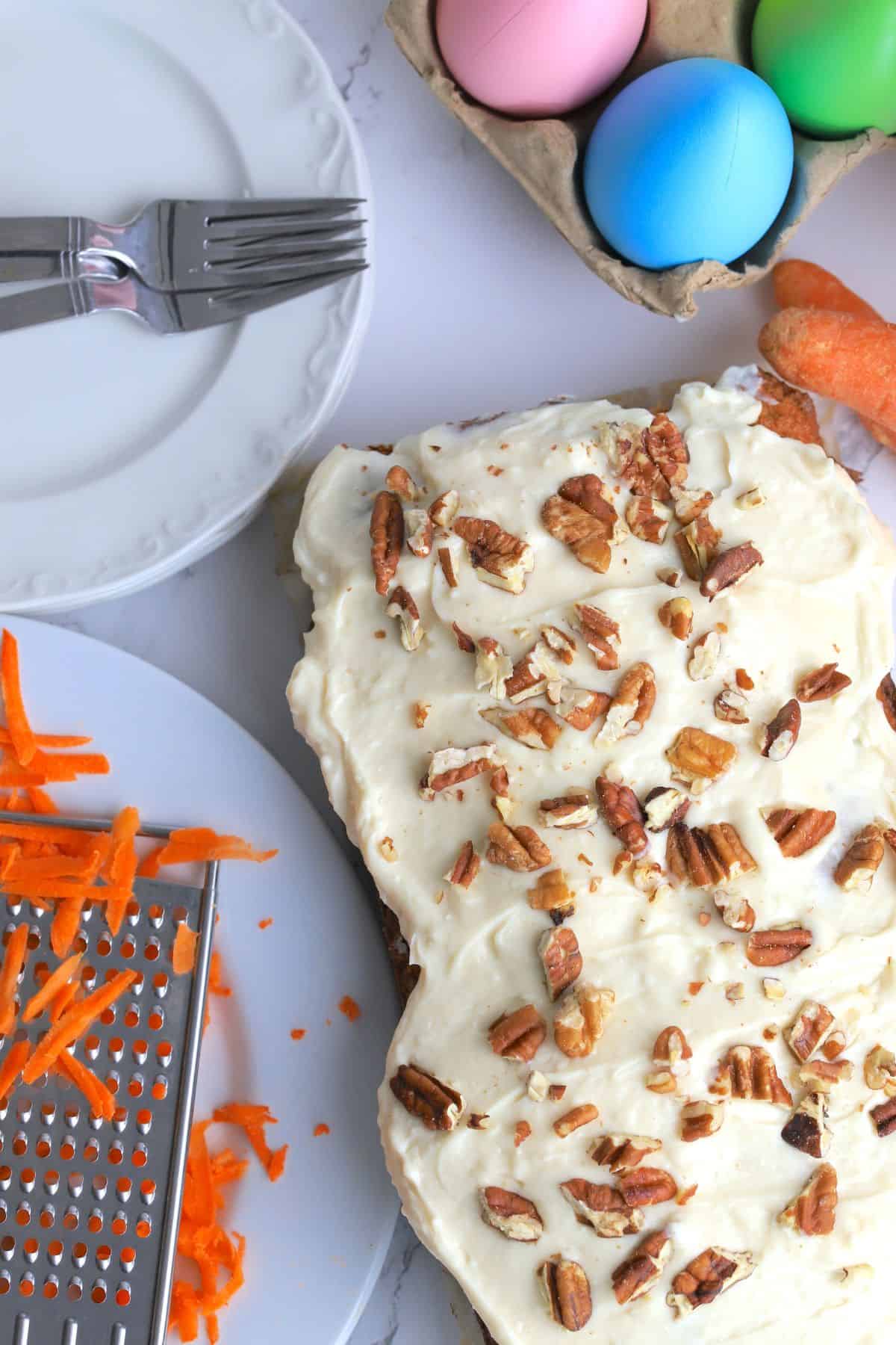 Sugar free carrot cake next to a coffee mug, dessert plates and shredded carrots with colored easter eggs.